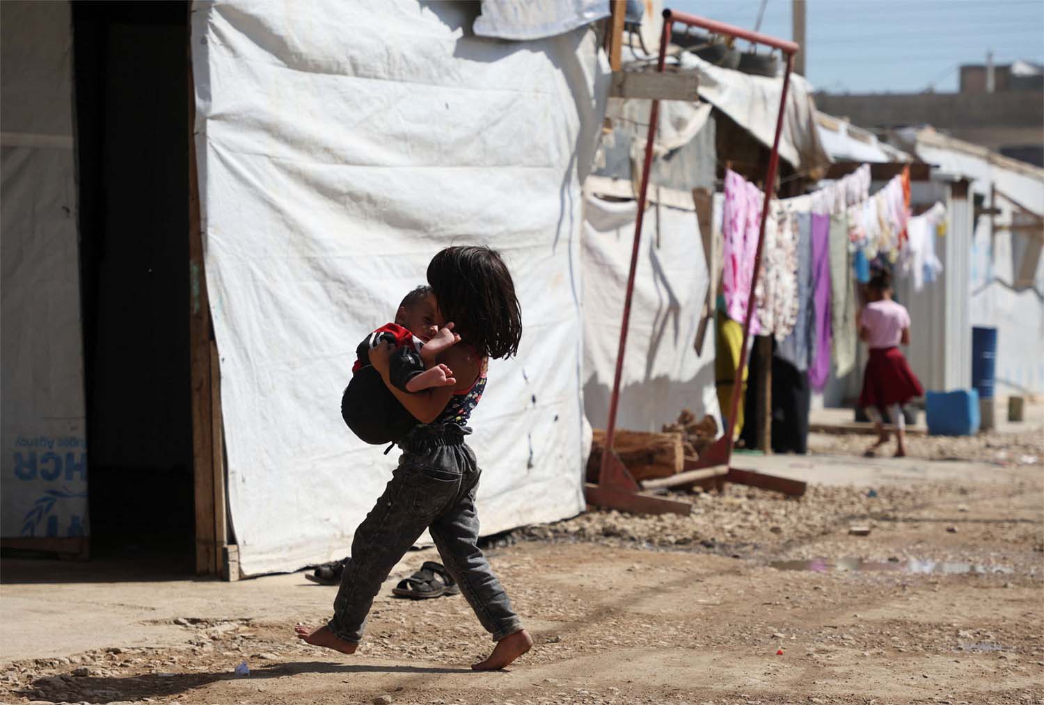 A Syrian refugee girl carries a child as she walks past tents at an informal camp in Bekaa Valley
