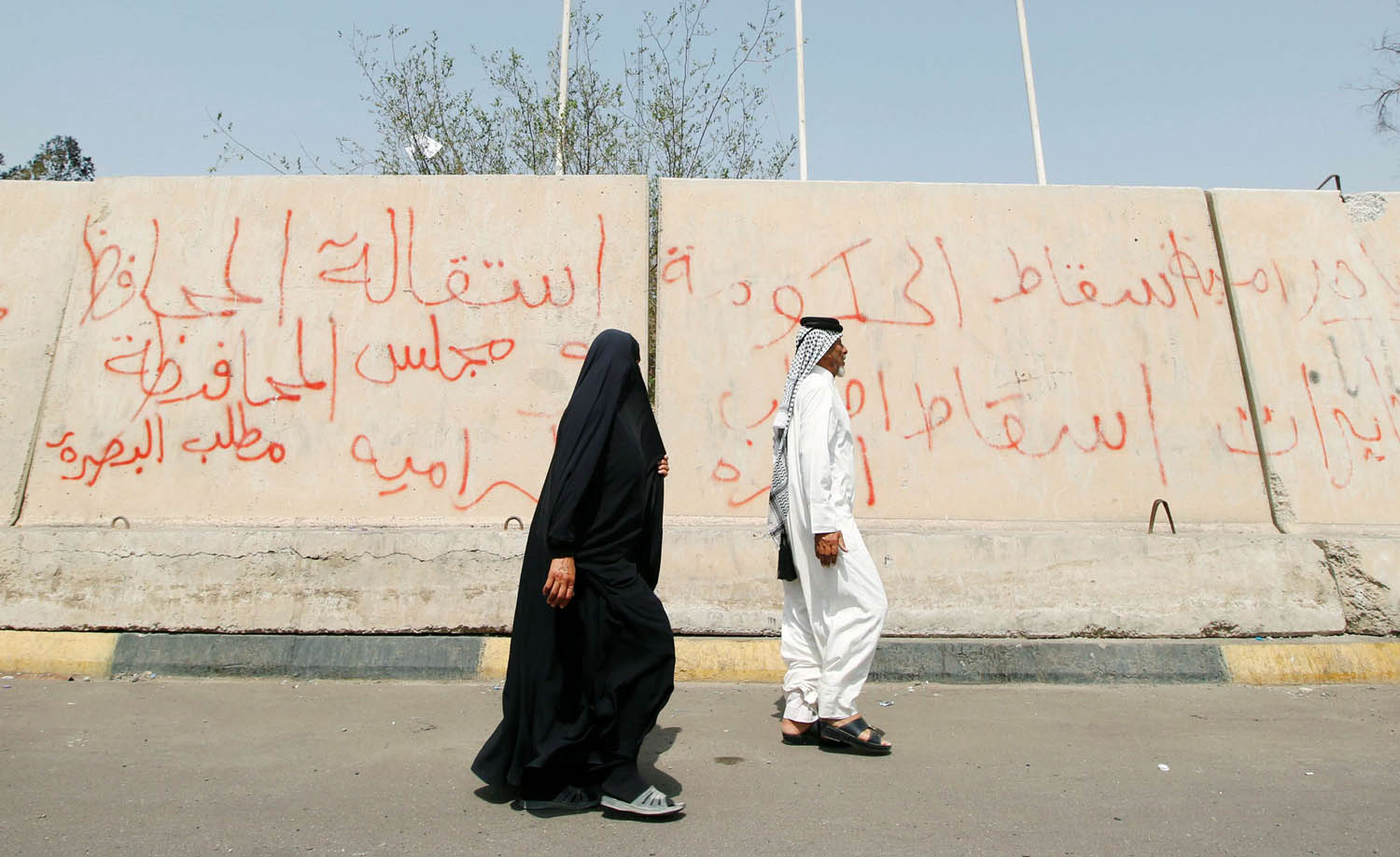 عراقي وعراقية يمران بجانب جدار يحمي منشأة حكومية في البصرة تلطخه الشعارات
