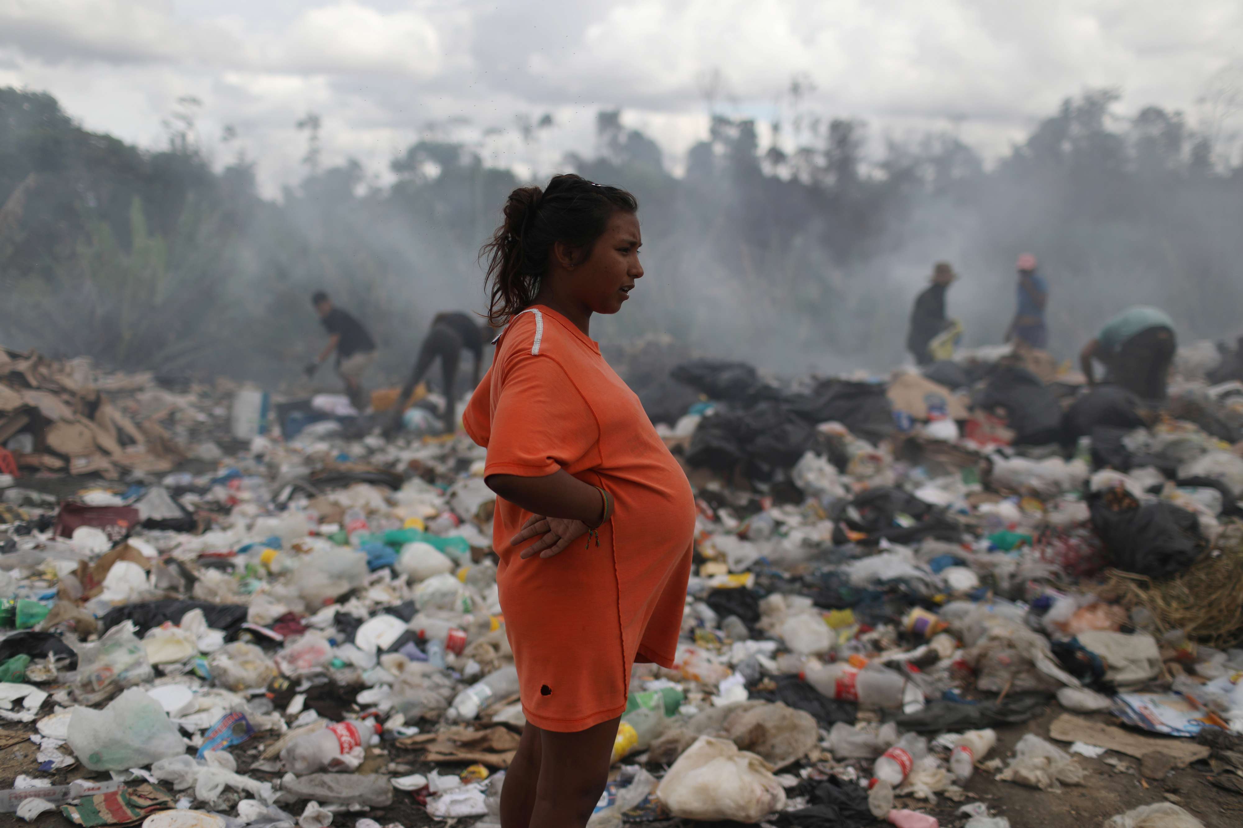 امرأة حامل تقف قرب مصب نفايات يتصاعد منه الدخان