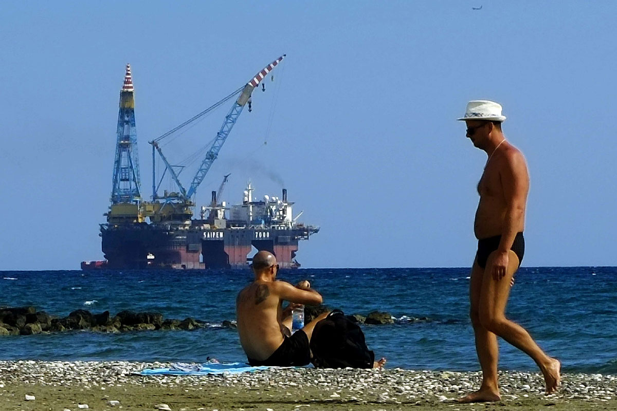 Men sit on a beach as a drilling platform is seen in the background near Larnaca