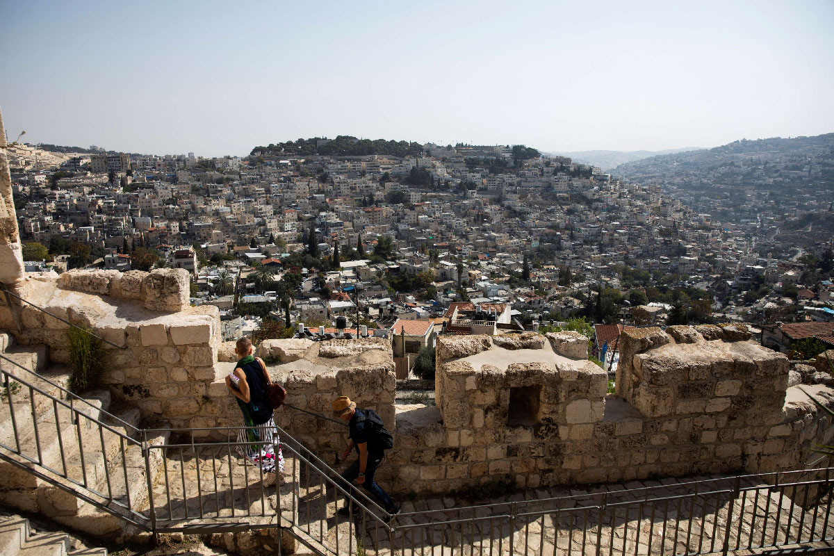 The Palestinian neighborhood of Silwan is seen in the background as people walk on a promenade on the surrounding walls of Jerusalem's Old City
