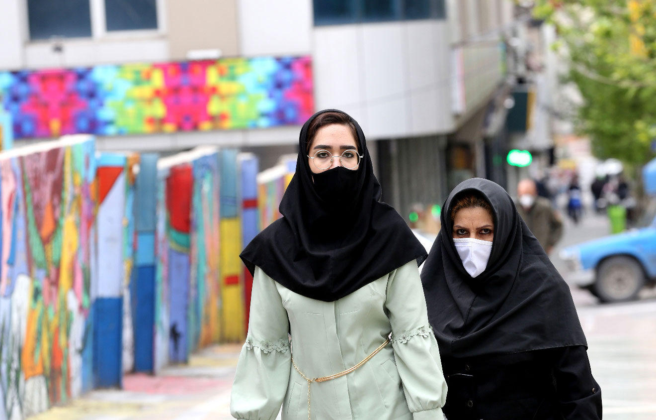 Iranians wearing protective mask walk past graffiti on a wall in Tehran