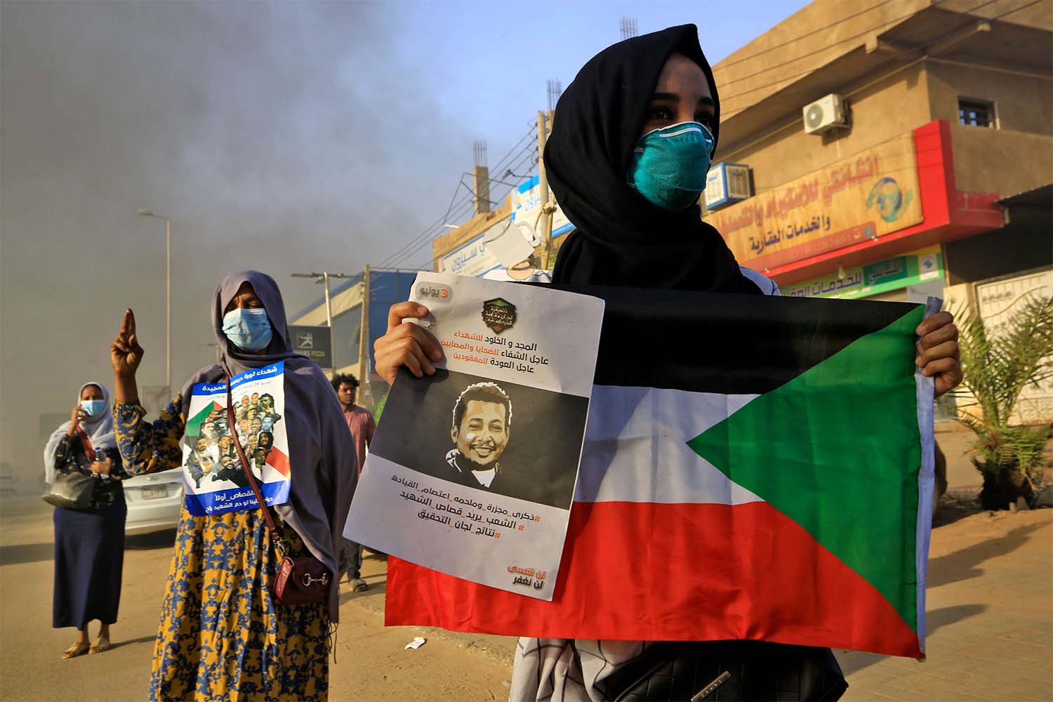 A mask-clad Sudanese woman holds a national flag along with a poster depicting the face of a slain protester