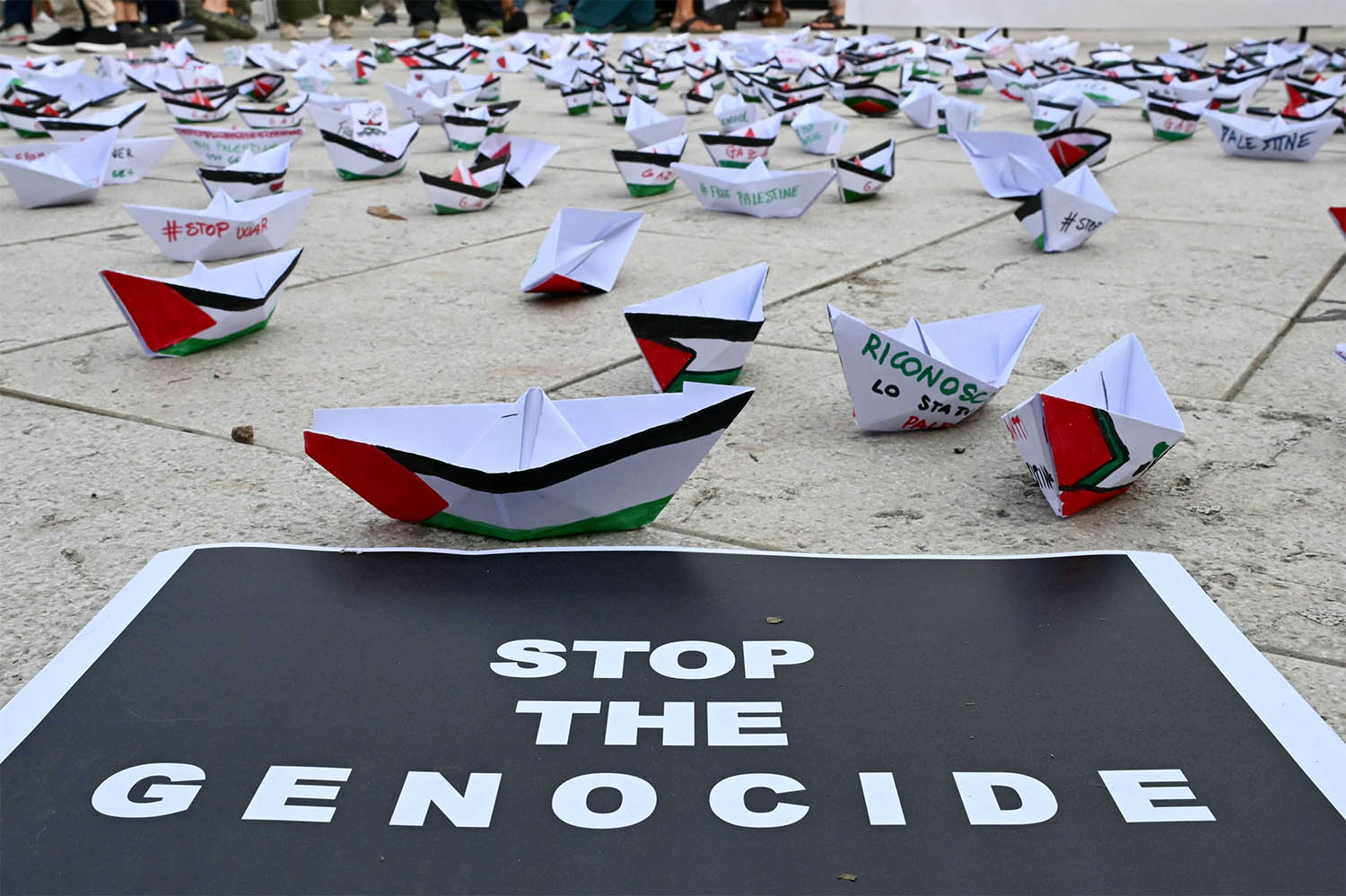 A flotilla of paper boats are laid on the ground near a poster reading Stop the Genocide during a demonstration in support of Gaza and Palestinian people at Venice Lido