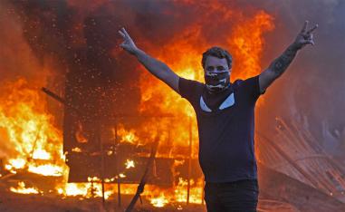A protester flashes the V for victory sign as a riot police vehicle burns behind him during protests in Baghdad