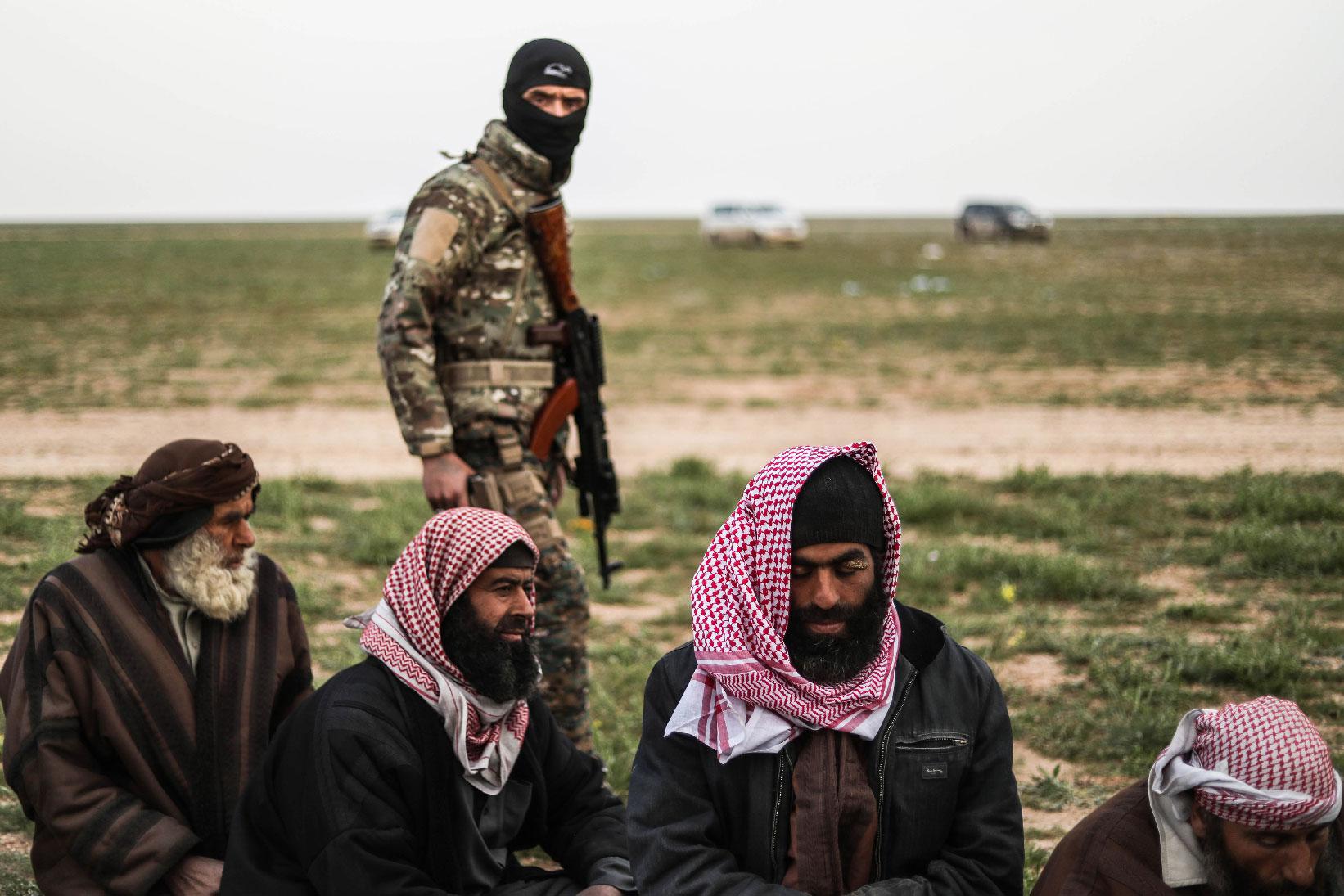 A fighter with the US-backed Syrian Democratic Forces (SDF) walks past men at a screening area for those who are evacuated from the Islamic State (IS) group's embattled holdout of Baghouz, during an operation to expel IS jihadists from the area, in the eastern Syrian province of Deir Ezzor, on February 26, 2019.