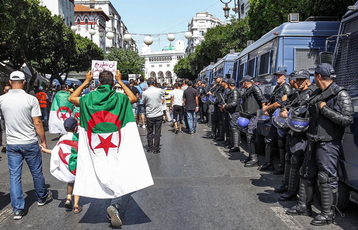 Protesters draped in Algerian national flags walk past riot police members