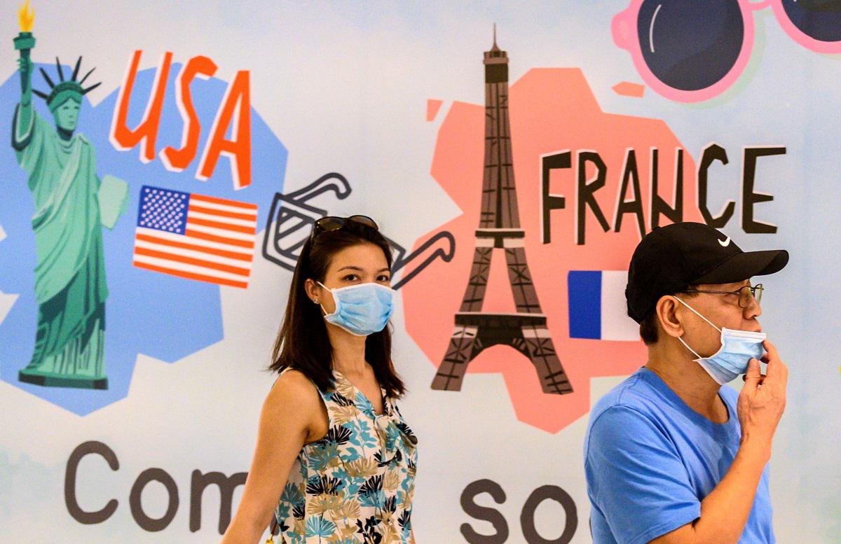 أشخاص يرتدون أقنعة واقية يمشون أمام لوحة إعلانية في بانكوك تظهر معالم سياحية عالمية