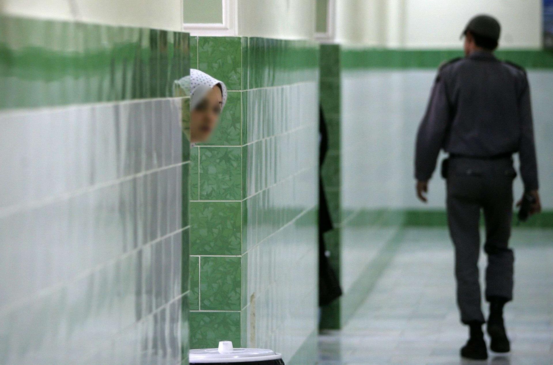 An Iranian inmate peers from behind a wall as a guard walks by