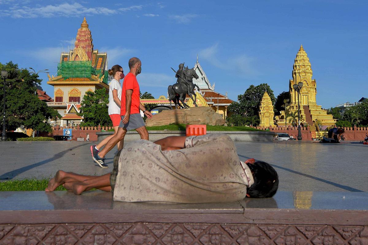 رجل بلا مأوى يتمدد على الأرض بينما يمشي زوجان يرتديان كمامات في بنوم بنه عاصمة كمبوديا