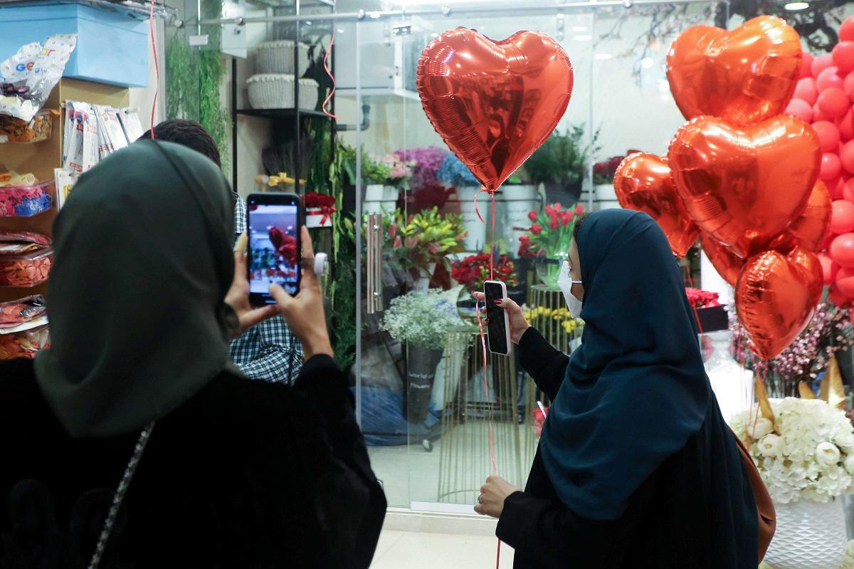 سعودية تلتقط صورة بهاتفها المحمول بينما تحمل امرأة أخرى بالونًا على شكل قلب أثناء تسوقها قبل عيد الحب في الرياض